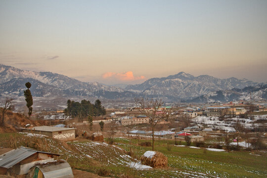 View of Chattar Plain with snow-covered mountains in the background, rural buildings, and agricultural fields under a soft sunset sky in Chattar Plain, Khyber Pakhtunkhwa, Pakistan.