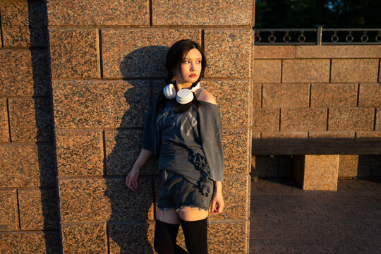 Woman with headphone stands by a city wall during summer evening