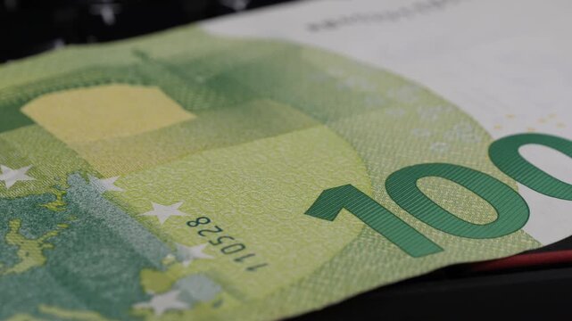 Close-up view of a 100 Euro banknote displayed on a black surface, revealing intricate details and textures, with a keyboard partially visible in the background enhancing the financial context