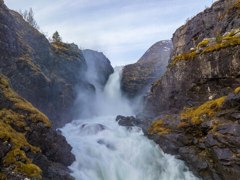 View of a powerful waterfall cascading between steep, moss-covered rocky cliffs with rushing white water in the foreground in Valldal, More og Romsdal, Norway.