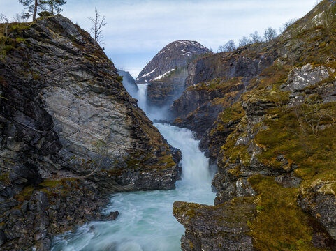 View of a powerful waterfall cascading through a narrow rocky gorge with moss-covered cliffs and a snow-capped mountain in the background in Valldal, More og Romsdal, Norway.