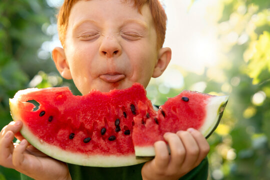 Playful red-haired boy eating watermelon in the garden