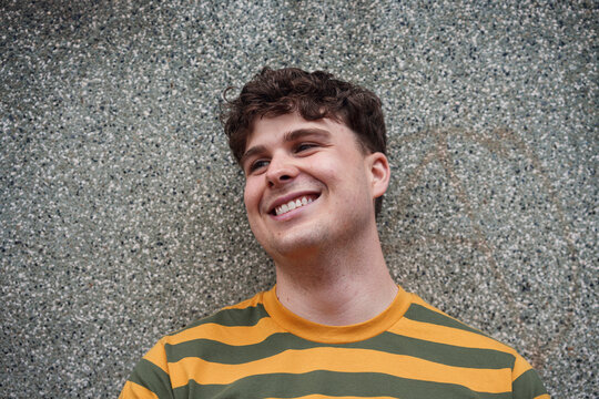 A smiling man leans against a textured wall outdoors