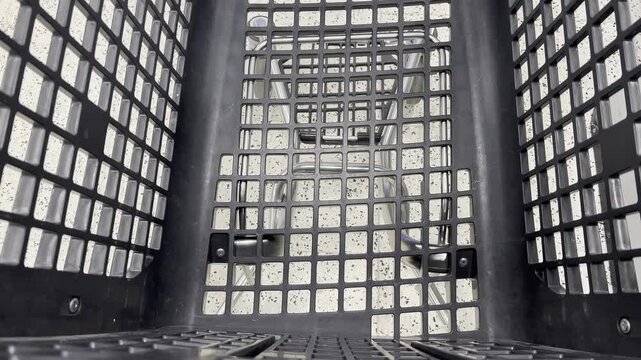 Black plastic shopping basket viewed from above, revealing a grid pattern and interior structure. The floor is visible, with a speckled design, enhancing the overall visual context