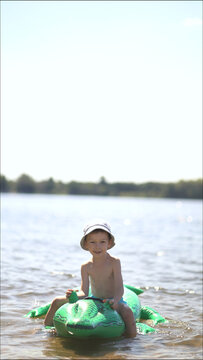 A happy young boy in a bucket hat sits smiling on a green inflatable crocodile float in a sunny lake, enjoying summer outdoor fun and childhood adventure.