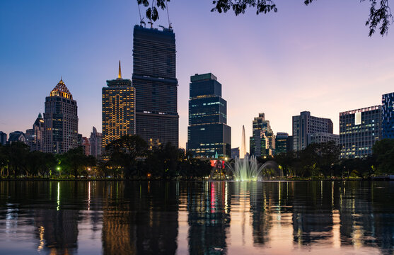 View of Lumphini Park with a fountain in the lake and the illuminated Bangkok skyline reflecting on the water at twilight in Bangkok, Thailand.