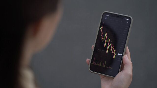 Woman trader closely monitors live Bitcoin price on her smartphone, analyzing red and green candlestick charts and volume for trading decisions.