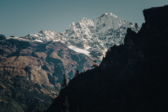 Majestic snow-covered mountain peaks rise above a dramatic Himalayan valley, creating a serene wilderness landscape with deep shadows, rugged terrain, forest and ample copy space.