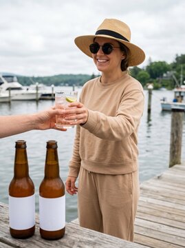 A smiling woman in a hat and sunglasses receives a refreshing drink on a wooden dock with boats in the background.