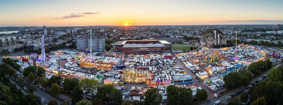 Hamburg, Germany - 24 August 2026: Aerial view of Hamburger Dom fairground, featuring various amusement rides, roller coasters, and stalls, with the city skyline in the background under a golden sky.