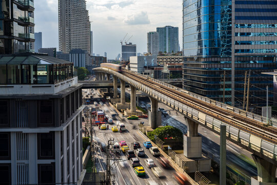 View of the BTS Skytrain tracks with a passing train above busy city traffic and modern skyscrapers in Bangkok, Thailand.