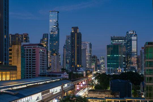 View of the Bangkok skyline at dusk featuring the iconic MahaNakhon building, illuminated skyscrapers, and the BTS Skytrain tracks seen from Surasak BTS, Bangkok, Thailand.