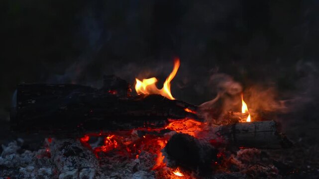 Seamless Looping Video of Bonfire Flames and Glowing Wood. Close-up seamless video loop of intensely glowing red embers, hot charcoal, and flickering flames on firewood logs in a dark outdoor setting.