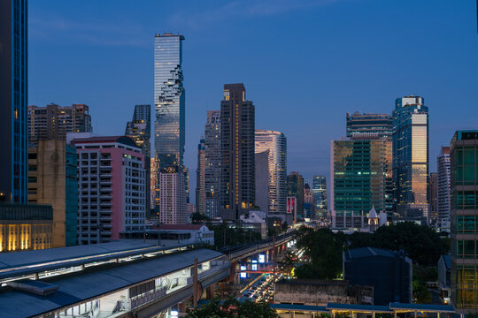 View of the Bangkok skyline at dusk featuring the iconic pixelated architecture of the King Power Mahanakhon building rising above urban high-rises and BTS Skytrain tracks in Bangkok, Thailand.