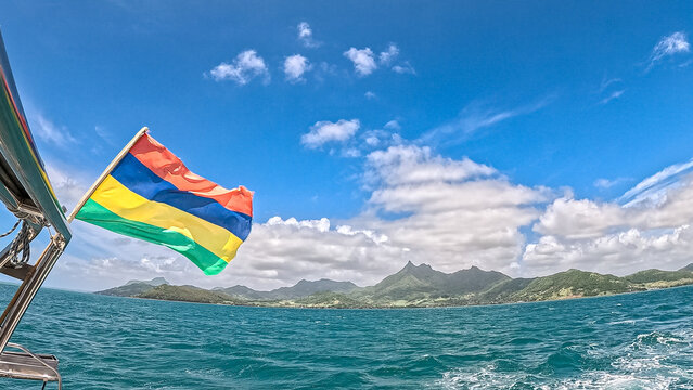 View of the national flag of Mauritius waving from a boat against a backdrop of tropical mountains and the Indian Ocean in Trou-aux-Biches, Pamplemousses District, Mauritius.
