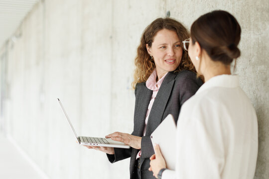 A businesswoman shows a laptop to a female colleague in the hallway