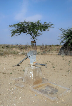 View of a metal hand-operated water pump standing in a dry, arid landscape with sparse vegetation and a small tree growing behind it in Nagarparkar, Sindh, Pakistan.