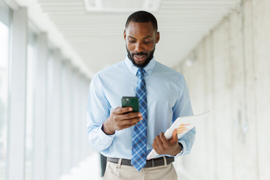 Businessman using smartphone and holding document in office corridor