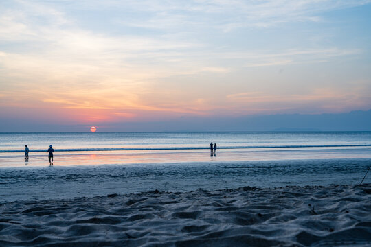 View of Ao Yai beach at sunset with people walking along the wet sand and calm ocean waves in Ko Phayam, Ranong, Thailand.