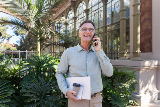 Smiling businessman talks on smartphone holding laptop outdoors