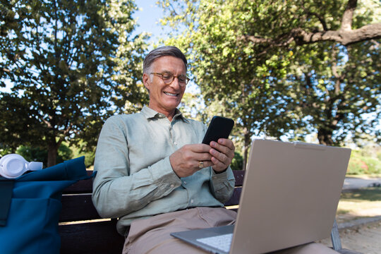 Smiling man uses smartphone and laptop on a park bench