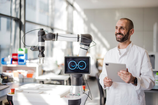 Smiling scientist holds a tablet next to a robot in a laboratory