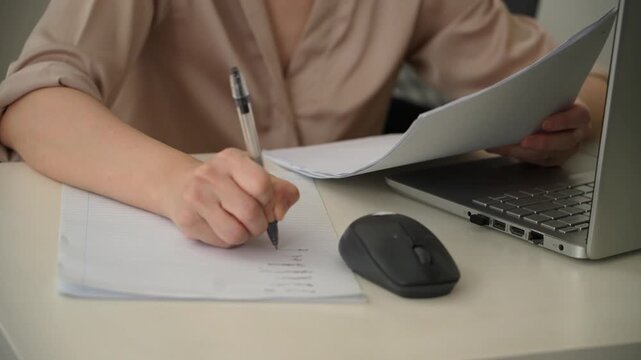 Close up footage of a woman writing notes on lined paper while consulting documents beside a laptop and wireless mouse at an office desk. Everyday planning, paperwork and administrative work.
