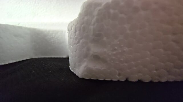 A close-up shot capturing the granular surface texture and cellular structure of a white styrofoam block against a dark fabric background with dramatic, soft side lighting. Perfect for material study.