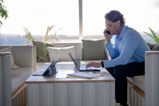Businessman talks on smartphone and works on laptop in sunny office