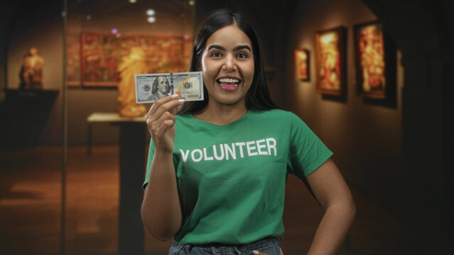 Volunteer woman holding a hundred dollar bill with hand raised in a museum smiling; charity joyful giving.