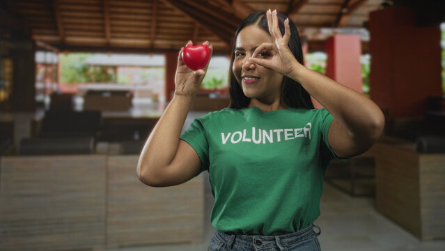 Woman volunteer holding a red heart and making an ok sign with hand in a wooden building lobby wearing volunteer tshirt; compassion charity community.
