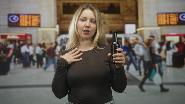 Blonde woman holding a beer bottle, hand to chest, standing amid a busy train station concourse with departure boards visible; tipsy confusion.