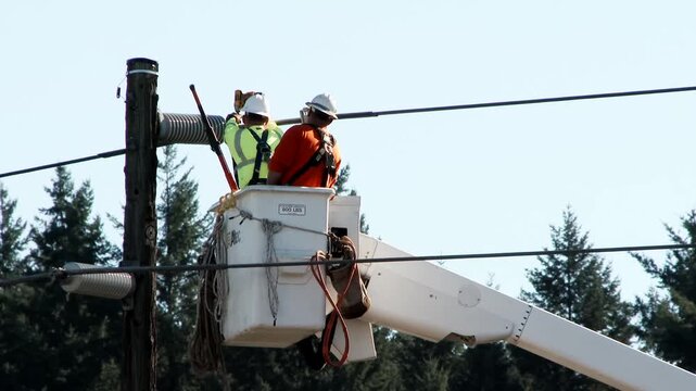 Two utility workers in elevated aerial lift performing overhead powerline maintenance beside weathered wooden utility pole under clear sky