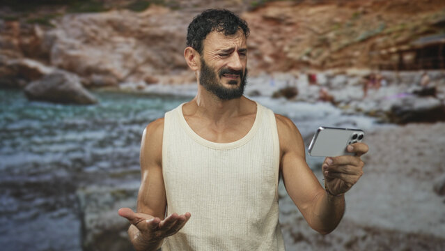 Man holds smartphone with bare forearm visible, grimacing while holding phone out at rocky beach backdrop in studio; frustration lost connection.
