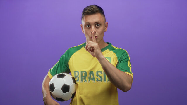 Man holding soccer ball, finger to lips for silence in studio wearing brazil jersey and winking; team pride.