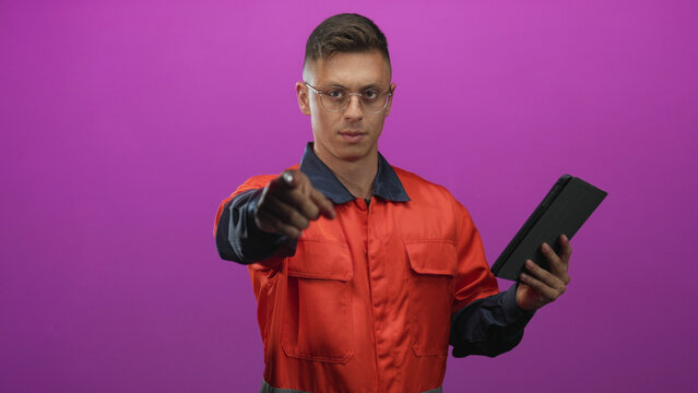 Man mechanic in orange coverall holds tablet and beckons with hand in studio; confidence readiness skill.