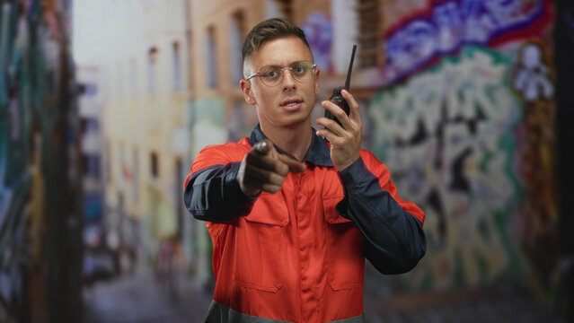 Man mechanic in orange coverall points finger at camera while holding radio in a graffiti street; vigilance duty.