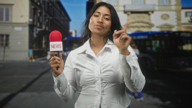 Woman reporter in white blouse holds red microphone labeled news and points index finger up on street with bus and historic building; confident reporting update.