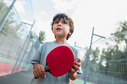 Young boy holding a red table tennis paddle outside on a court