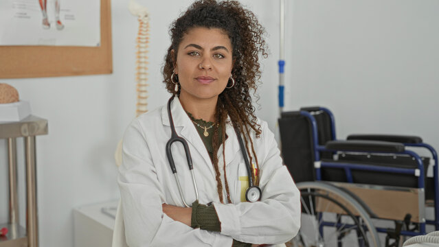 Woman doctor wearing white coat and stethoscope, standing with arms crossed before wheelchair and spine model in clinic building; compassion care confident.