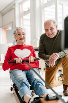 A male teacher cheers on a smiling woman on a rowing machine indoors
