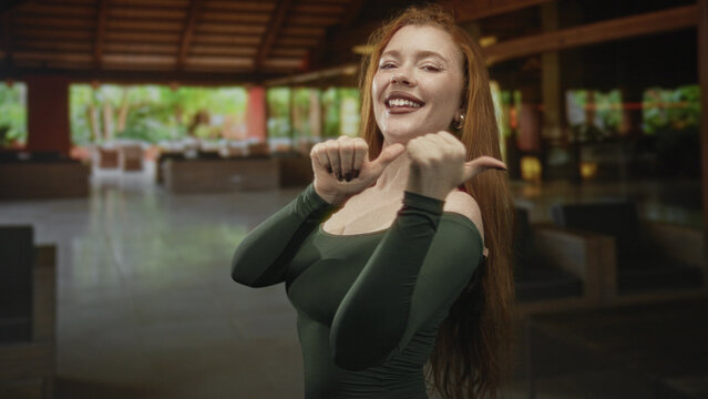 Redhead young woman smiling, showing thumbs up and bare shoulder inside a building lobby, wearing green off shoulder dress; joy playful.