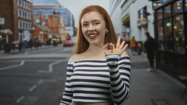 Redhead woman in striped off shoulder crop top making ok sign with right hand and bare shoulder visible on street; approval confidence.