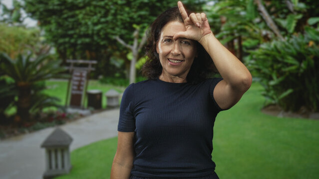 Woman making l gesture on forehead with index finger, smiling and tilting head beside stone lantern and garden path in a park; playful teasing.