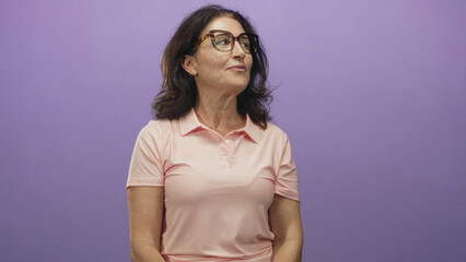 Obraz premium Middle aged hispanic woman in pink polo wearing glasses turns head to the right and smiles while seated against a purple studio backdrop; contentment.