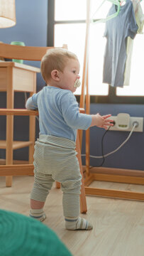 Toddler boy with pacifier holds wooden chair rail while standing and reaching in sunlit nursery by window with hanging clothes; curiosity.