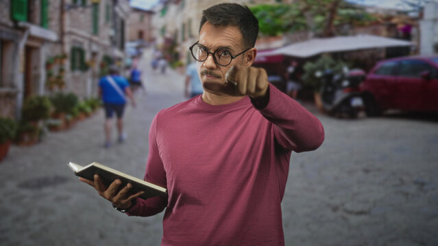 Hispanic man holding an open book while adjusting his glasses and pointing, standing on a cobblestone street lined with shops and planters; thoughtful study.