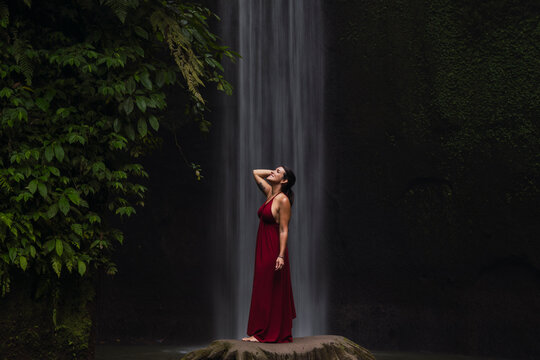 A woman in a red dress enjoys a waterfall in the Bali jungle