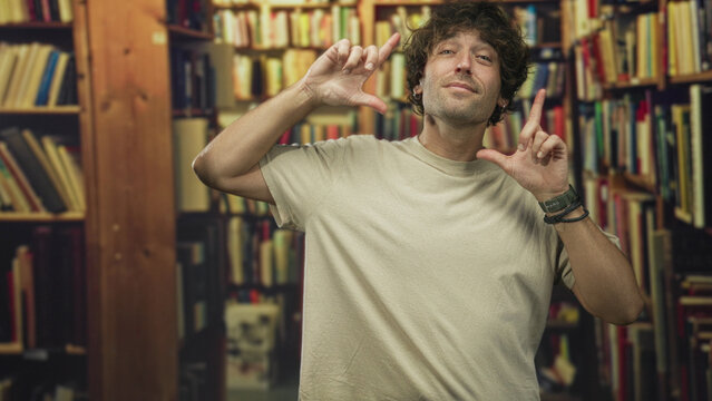 Young hispanic man framing face with fingers in front of bookshelves inside a building; playful confidence.