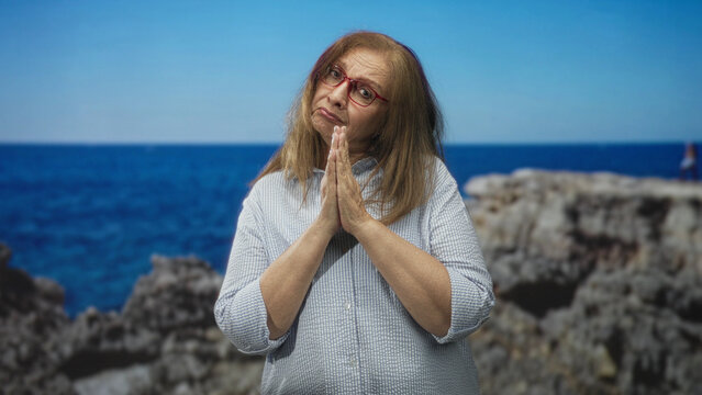 Woman with hands pressed together in prayer pose at studio seaside backdrop wearing red glasses and striped shirt; hopeful reflection.